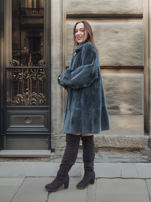 Model leans against stone wall in The Still Hour sheared muskrat fur coat in deep teal, styled for elegant winter outerwear fashion.