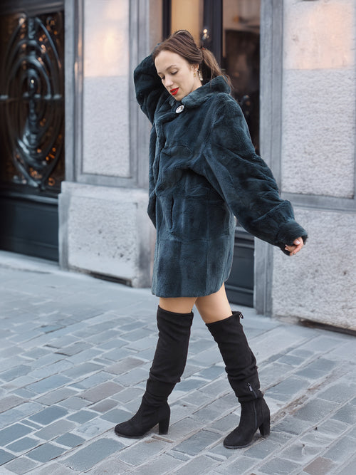Elegant and warm, The Still Hour sheared muskrat fur coat in deep teal flows effortlessly as the model walks in black knee-high boots.