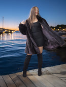 Woman in a black outfit with a fur coat standing on a dock by water at dusk.