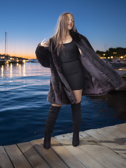 Woman in a black outfit with a fur coat standing on a dock by water at dusk.