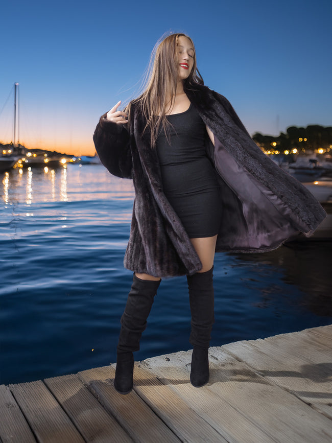 Woman in a black outfit with a fur coat standing on a dock by water at dusk.