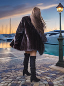 Person wearing a fur coat and knee-high boots standing by a marina at sunset.