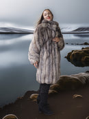 Person wearing a fur coat standing by a reflective body of water.