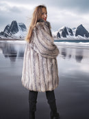 Woman wearing a fur coat standing in front of a mountainous landscape with water.