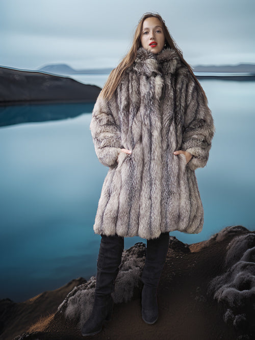 Person wearing a fur coat standing on a rocky landscape with water in the background