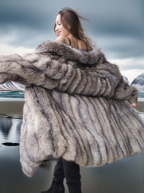 Person wearing a fur coat standing by a body of water with mountains in the background