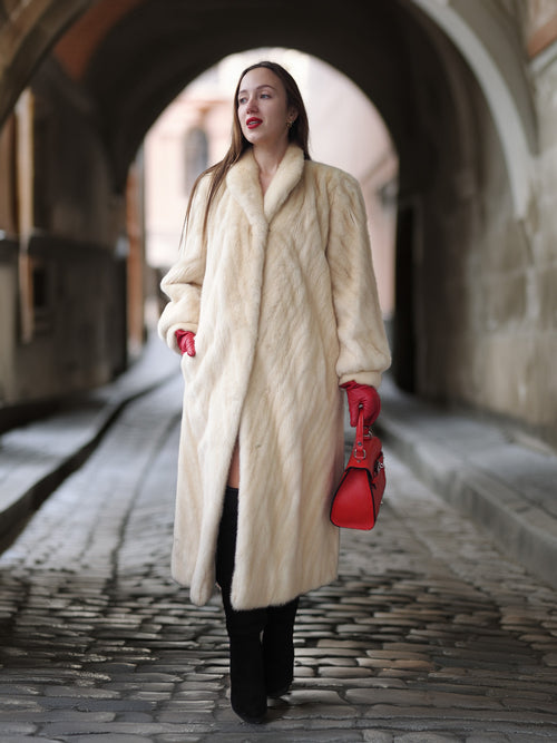 Woman in a long cream coat and red gloves holding a red bag on a stone street.