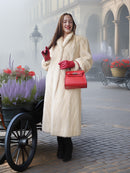 Woman in a long cream coat and red gloves holding a red handbag in an outdoor setting with flowers and a carriage.