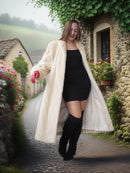 Woman in a long cream coat standing in a quaint village setting with stone buildings and flowers.