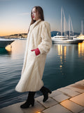 Woman in a long white fur coat standing by a waterfront with yachts in the background.