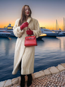 Woman in a fur coat and red gloves holding a red handbag by a marina at sunset.