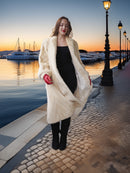 Woman in a long cream fur coat standing by a waterfront at sunset.