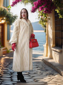 Woman in a long cream coat and red gloves holding a red handbag on a street with flowers.