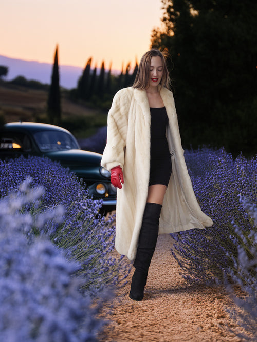 Woman in a long cream coat and black dress walking through lavender fields at sunset.