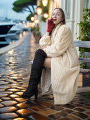 Woman in a long cream coat sitting on a bench with blurred lights and plants in the background