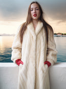 Woman in a long white fur coat and red gloves standing by a waterfront at sunset.