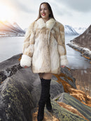 Woman in a fur coat standing on a rocky landscape with mountains and water in the background