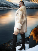 Woman in a fur coat standing by a lake with mountains in the background