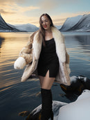 Woman in a fur coat standing by a lake with mountains in the background