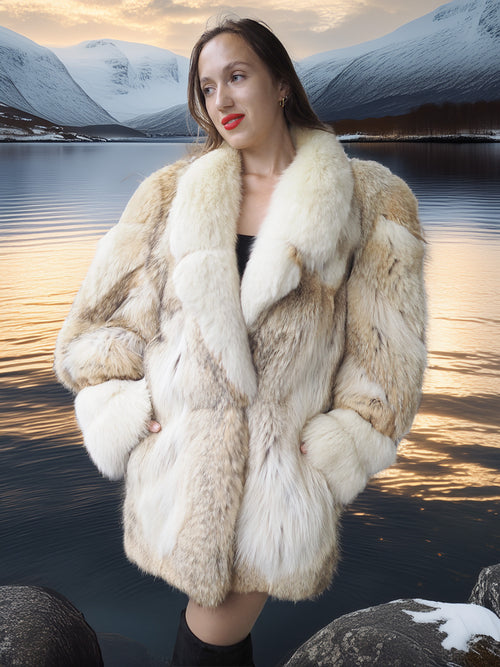Woman wearing a fur coat standing by a lake with mountains in the background