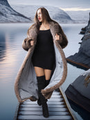 Woman in a long fur coat standing on a dock by a lake with mountains in the background