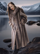 Woman in a fur coat standing by a lake with mountains in the background