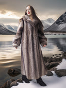 Person wearing a fur coat standing in a snowy landscape with mountains and water.