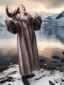 Woman in a fur coat standing by a lake with mountains in the background