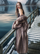 Woman in a fur coat standing on a snowy dock by a lake with mountains in the background