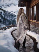 Woman in a fur coat standing in front of a wooden cabin with snow-covered mountains in the background