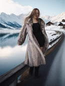 Woman in a fur coat standing by a snowy landscape with mountains and a lake.