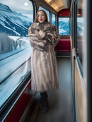 Woman in a fur coat standing inside a train with snowy mountain scenery outside.