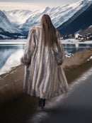 Person in a fur coat standing by a lake with snow-covered mountains in the background