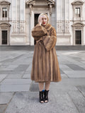 Elegant woman wearing long brown fur coat stands in front of ornate architectural building.