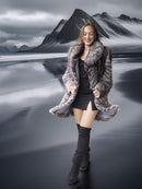 Woman in a fur coat standing in front of a mountainous landscape with a reflective lake.