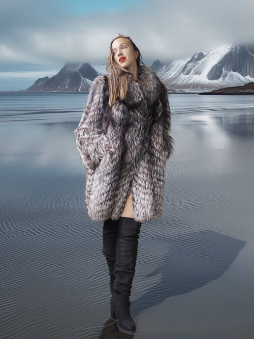 Woman in a fur coat standing on a reflective water surface with mountains in the background