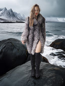 Woman in a fur coat and knee-high boots standing on a rocky shore with mountains in the background
