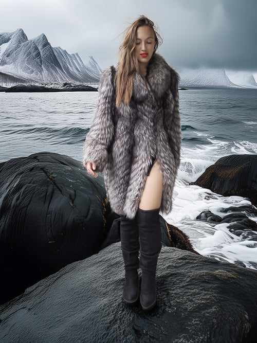 Woman in a fur coat and knee-high boots standing on a rocky shore with mountains in the background