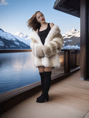 Woman in a fur coat and black boots standing by a window with a mountain view.