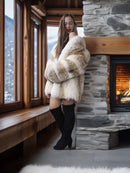 Woman in a fur coat standing in a cozy room with a fireplace and large windows.