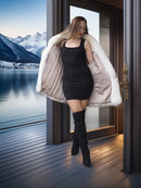 Woman in a black dress and white fur coat standing on a wooden deck with mountains and water in the background