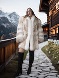 Woman in a fur coat and knee-high boots standing in front of a wooden cabin with mountains in the background.