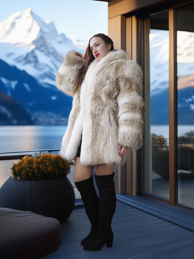 Woman in a fur coat standing in front of a scenic window with mountains and water.