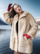 Woman in a fur coat and red gloves standing by a lake with mountains in the background