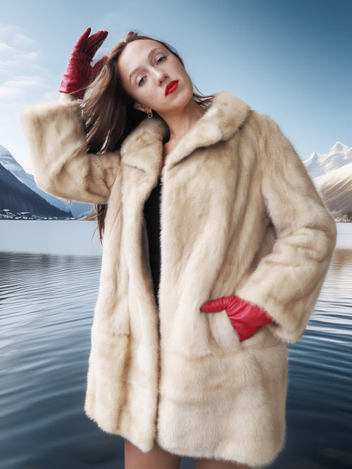 Woman in a fur coat and red gloves standing by a lake with mountains in the background
