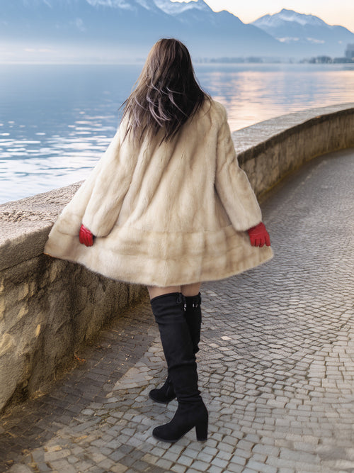 Person wearing a fur coat and black boots standing by a lakeside with mountains in the background