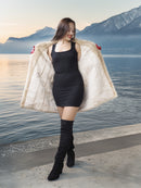 Woman in a black dress and knee-high boots with a fur coat by a lake with mountains in the background