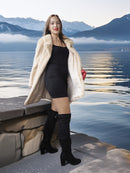 Woman in a fur coat standing by a lake with mountains in the background