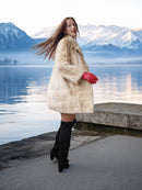 Woman in a fur coat and red gloves standing by a lake with mountains in the background