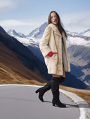 Woman in a fur coat and black boots standing on a mountain road with snow-capped mountains in the background